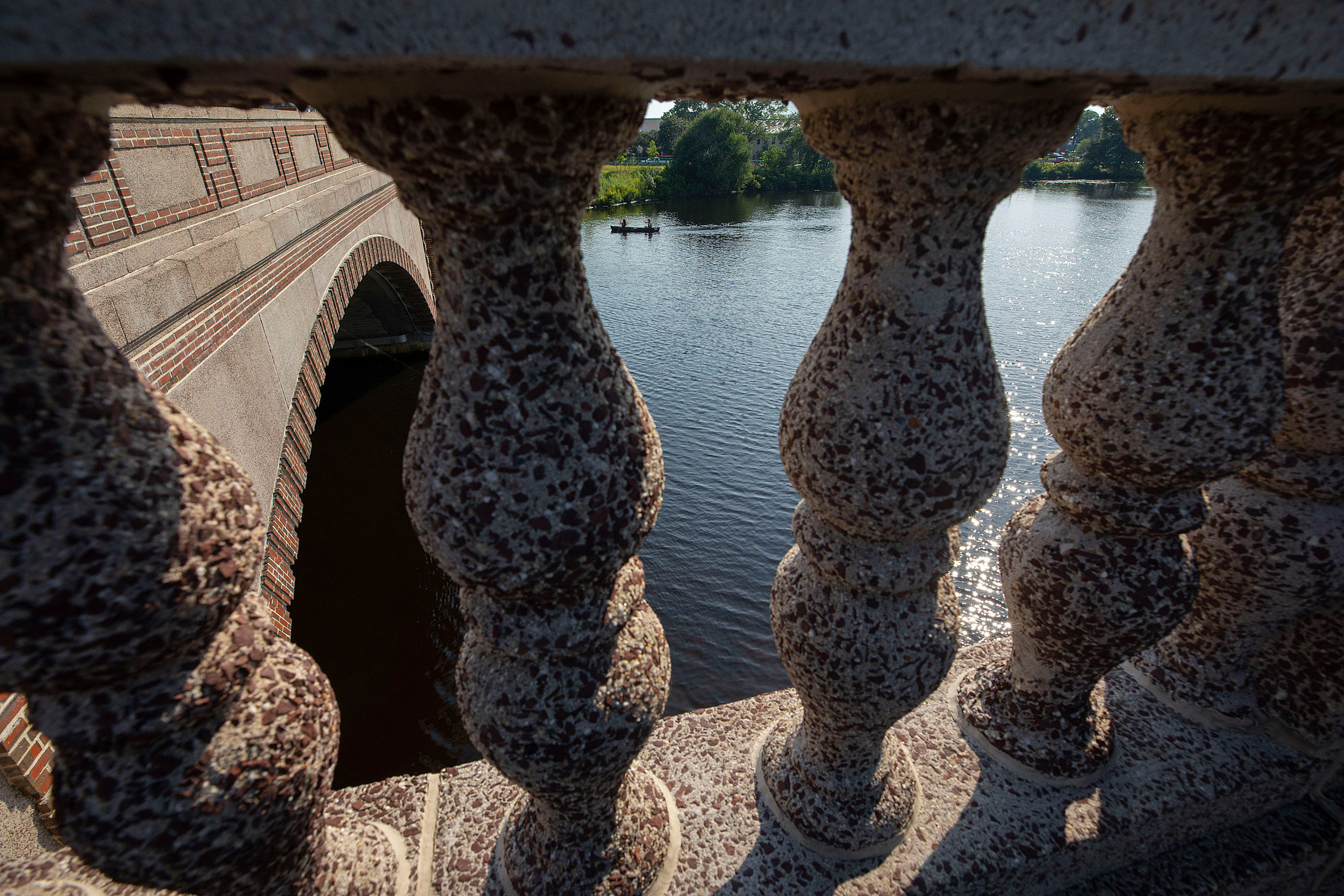 Looking through columns on a bridge looking over the Charles River at two people rowing a canoe.