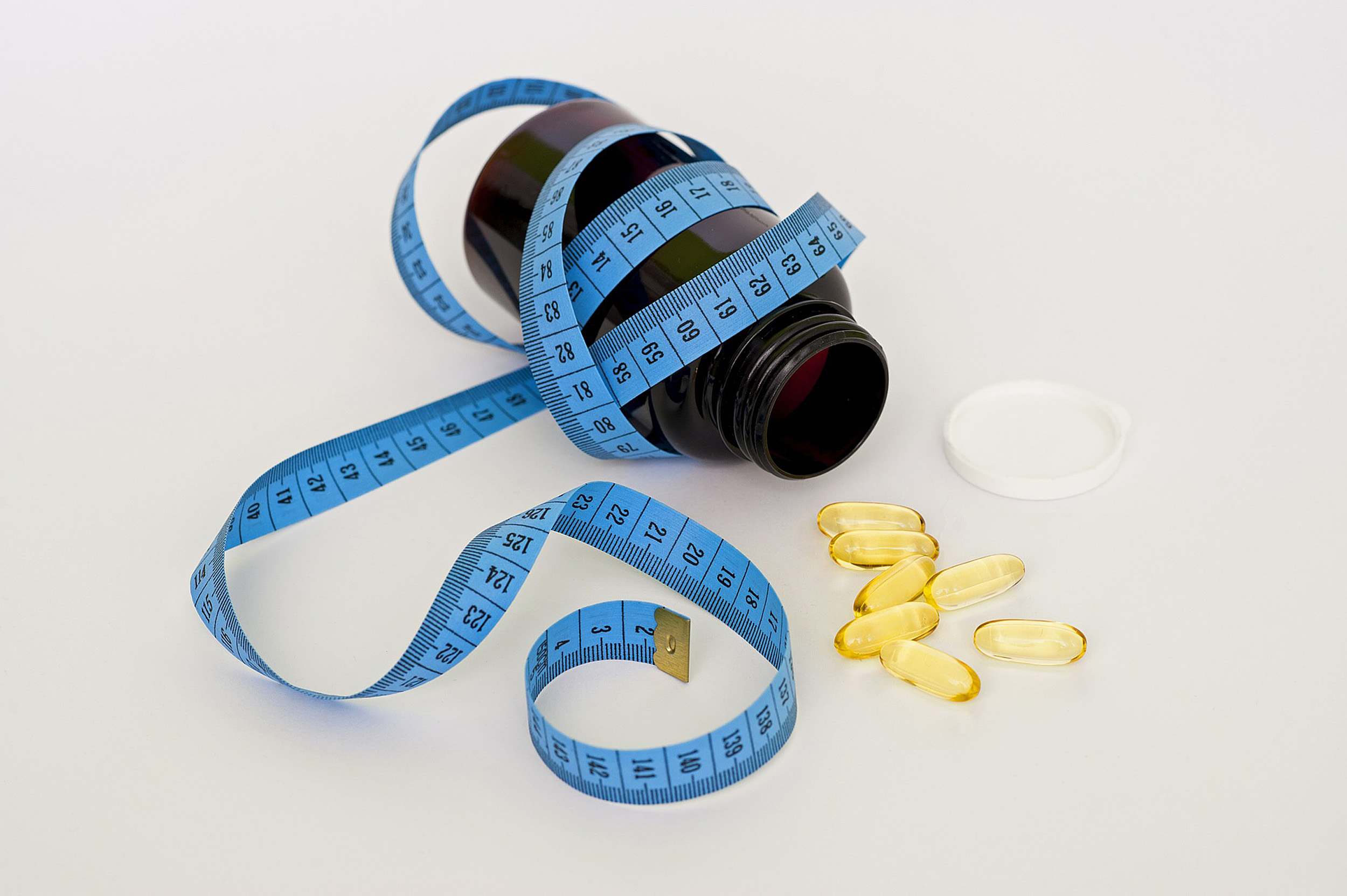 A blue measuring tape wrapped around a medicine bottle, with loose pills scattered around the open lid.