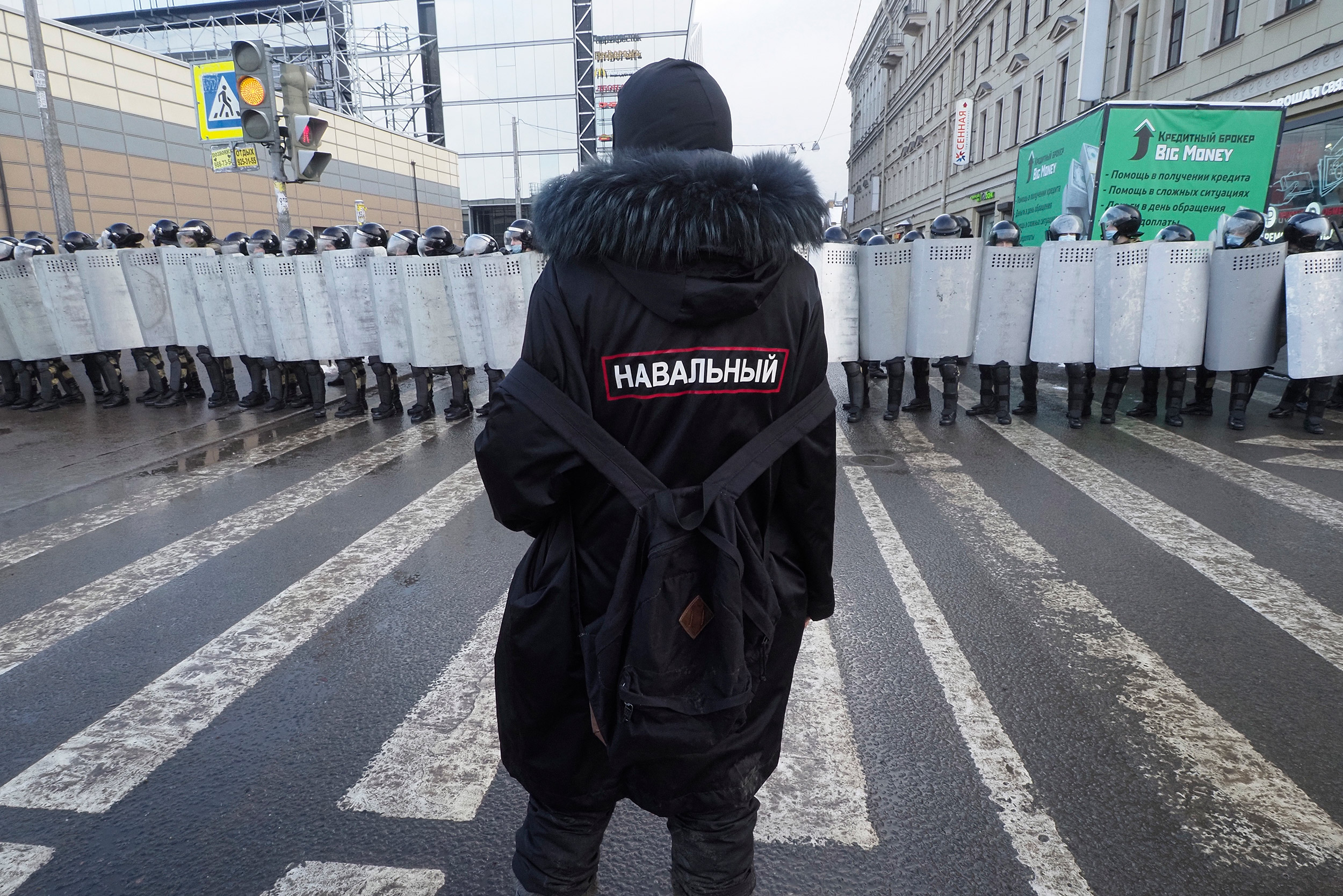 A man with a sign 'Navalny' on his back stands in front of riot policeme.