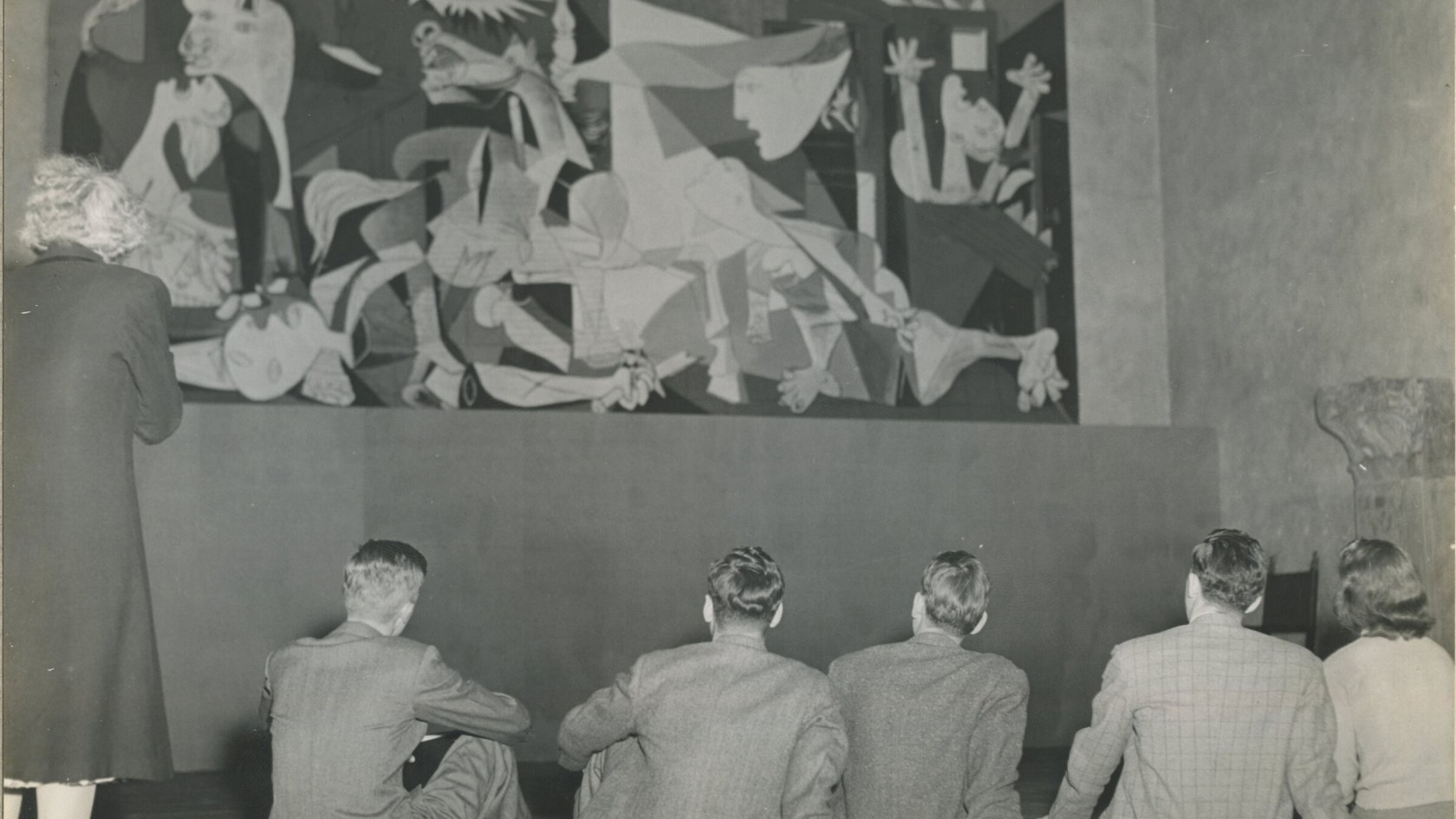One woman standing and four men sitting in from of the oil painting Guernica by Pablo Picasso.