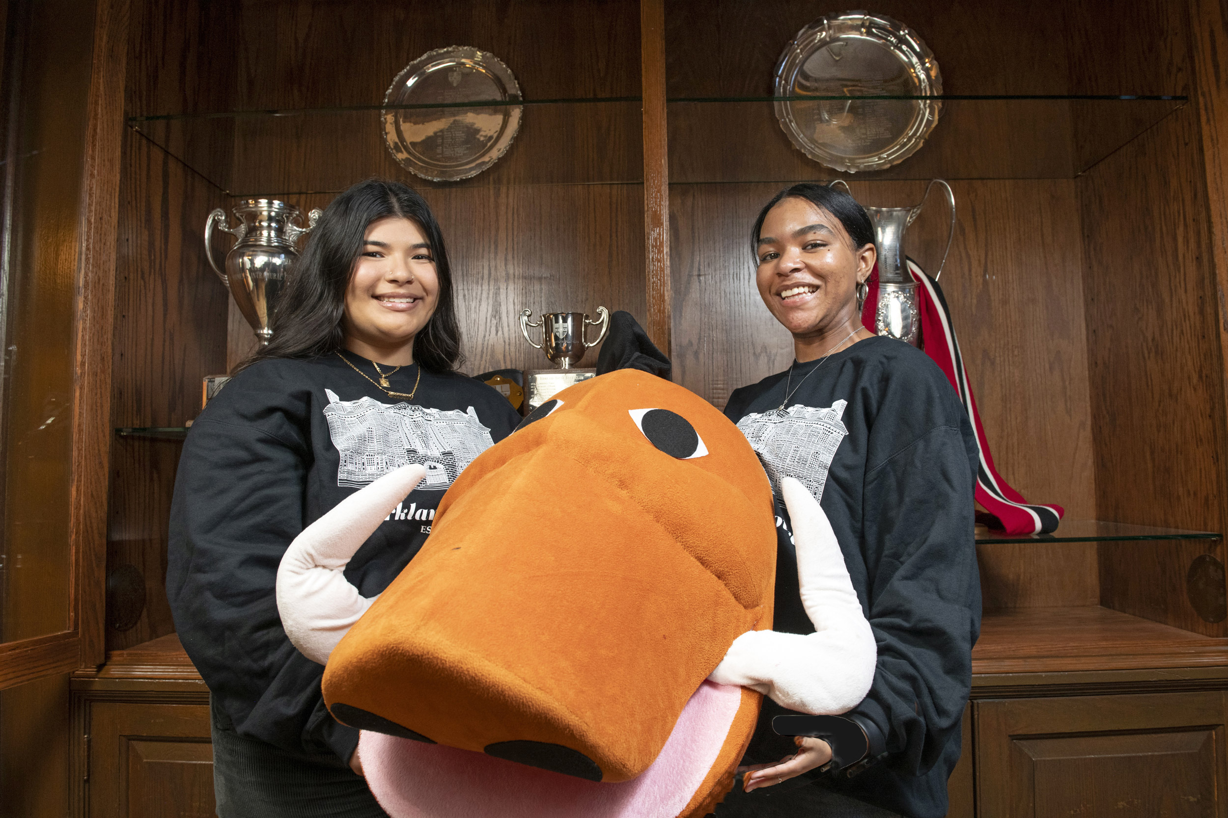 Giselle Chiprez (left) and Nena King with their boar mascot costume in front of a trophy case in the Kirkland House Junior Common Room.