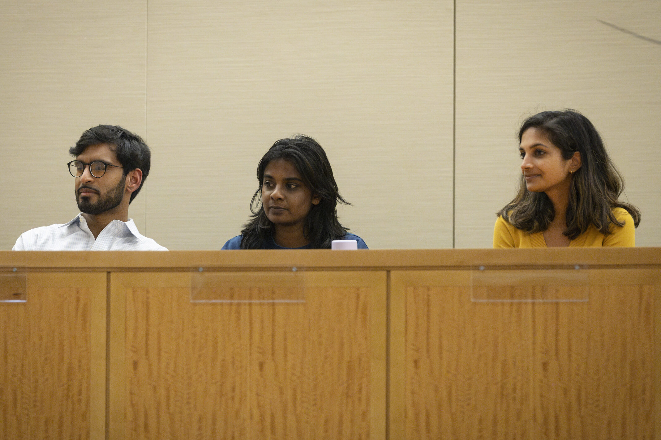 Three students listening to the speakers while sitting behind a wooden desk.