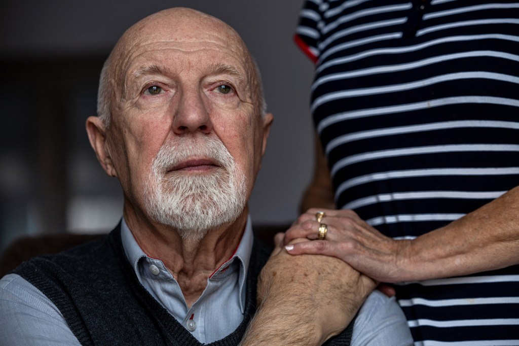 Portrait of a senior man with spouse's hand on his shoulder.