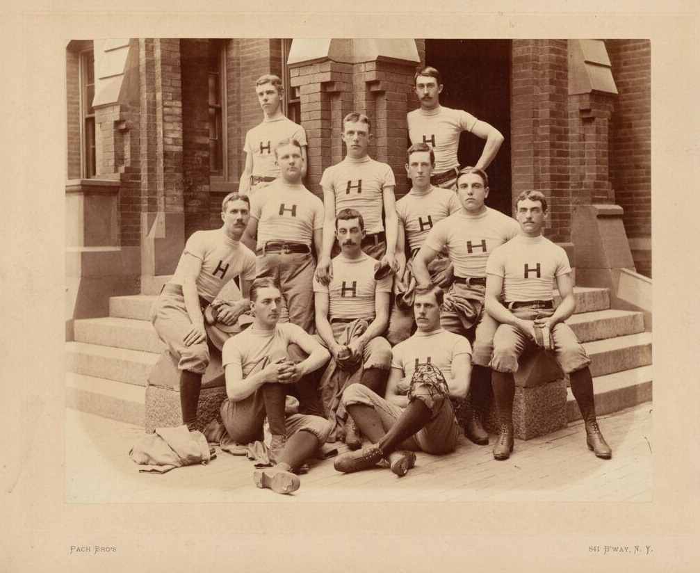 The 1884 Harvard baseball team poses by a brick building, wearing shirts with "H" on them