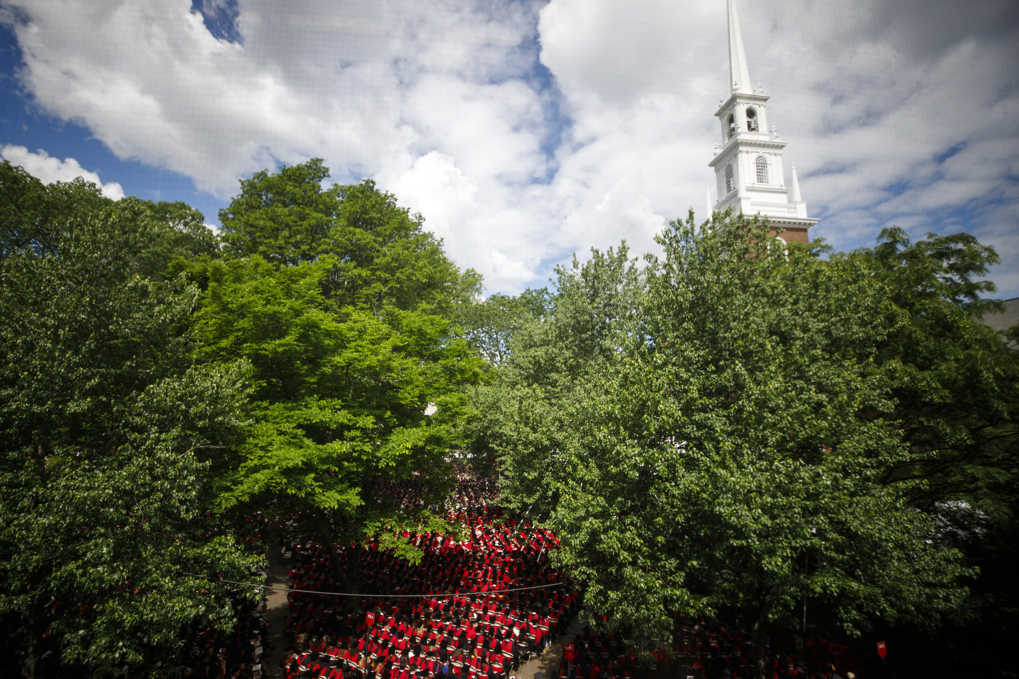 Graduates are seen through the windows of Sever Hall sitting in Tercentenary Theatre with the steeple of Memorial Church rising into the sky.