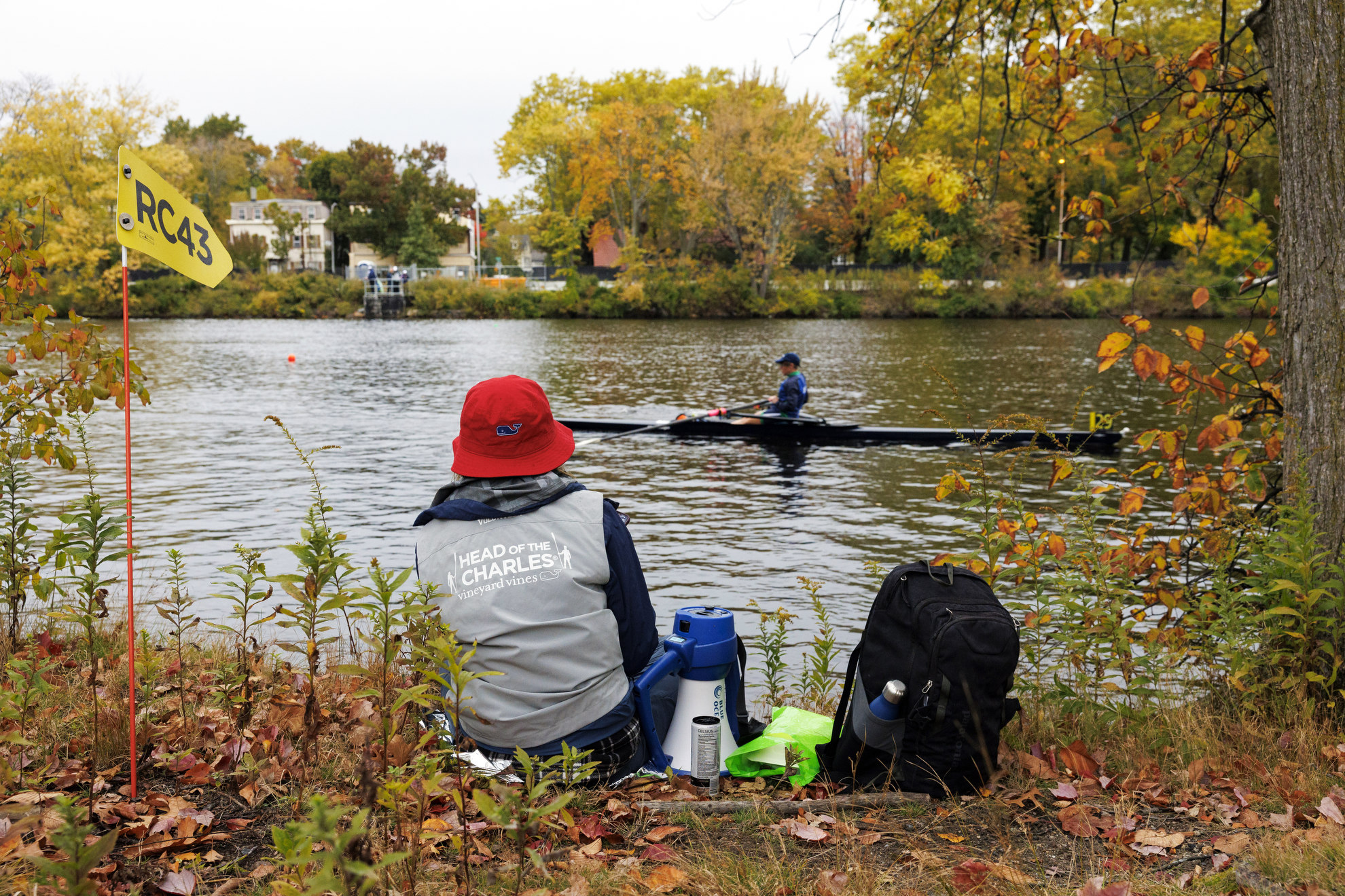 A Head of the Charles staffer sits on the banks of the Charles with their megaphone, watching a rower pass by.