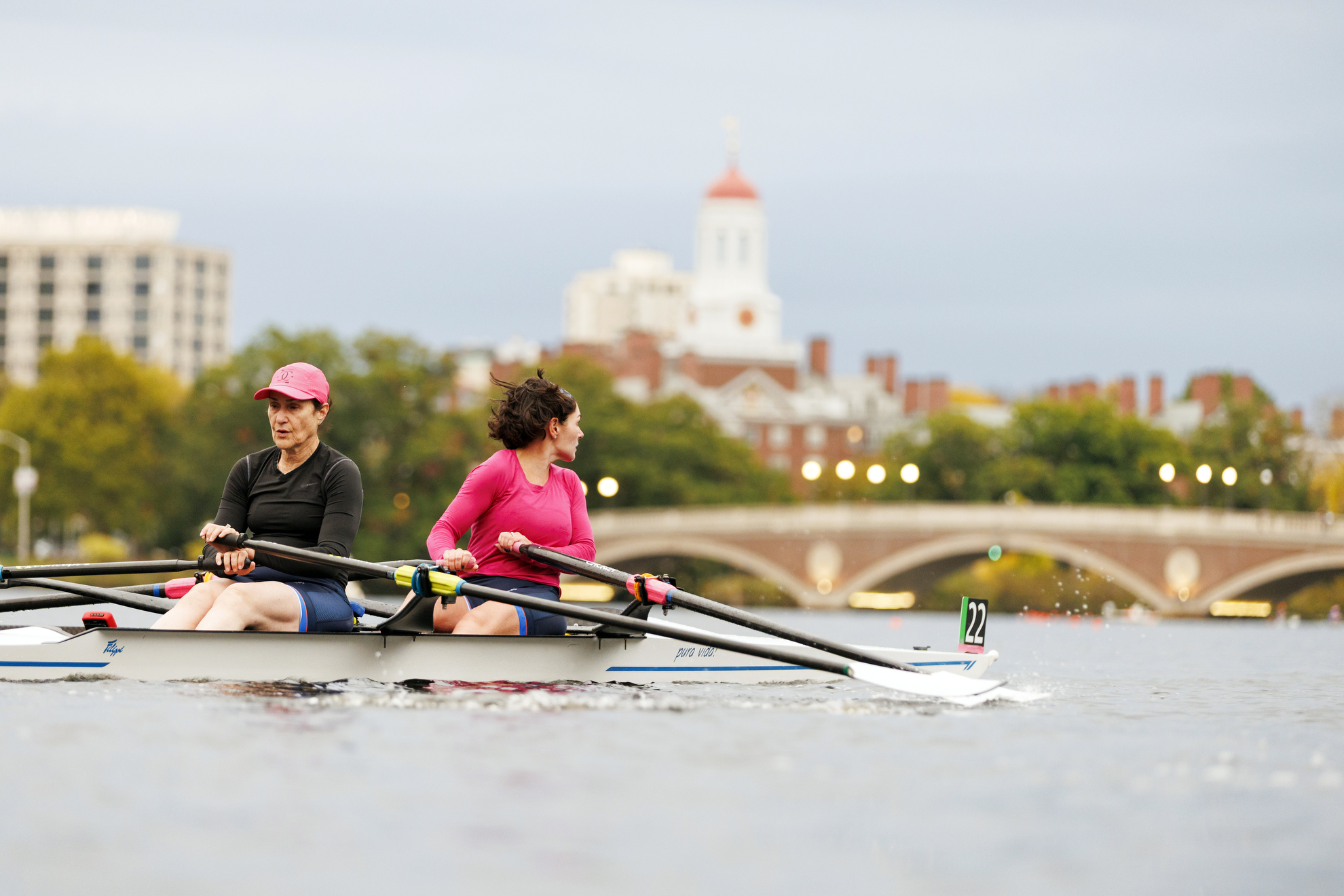 Two rowers head towards the John W. Weeks Footbridge with Dunster House visible in the background.
